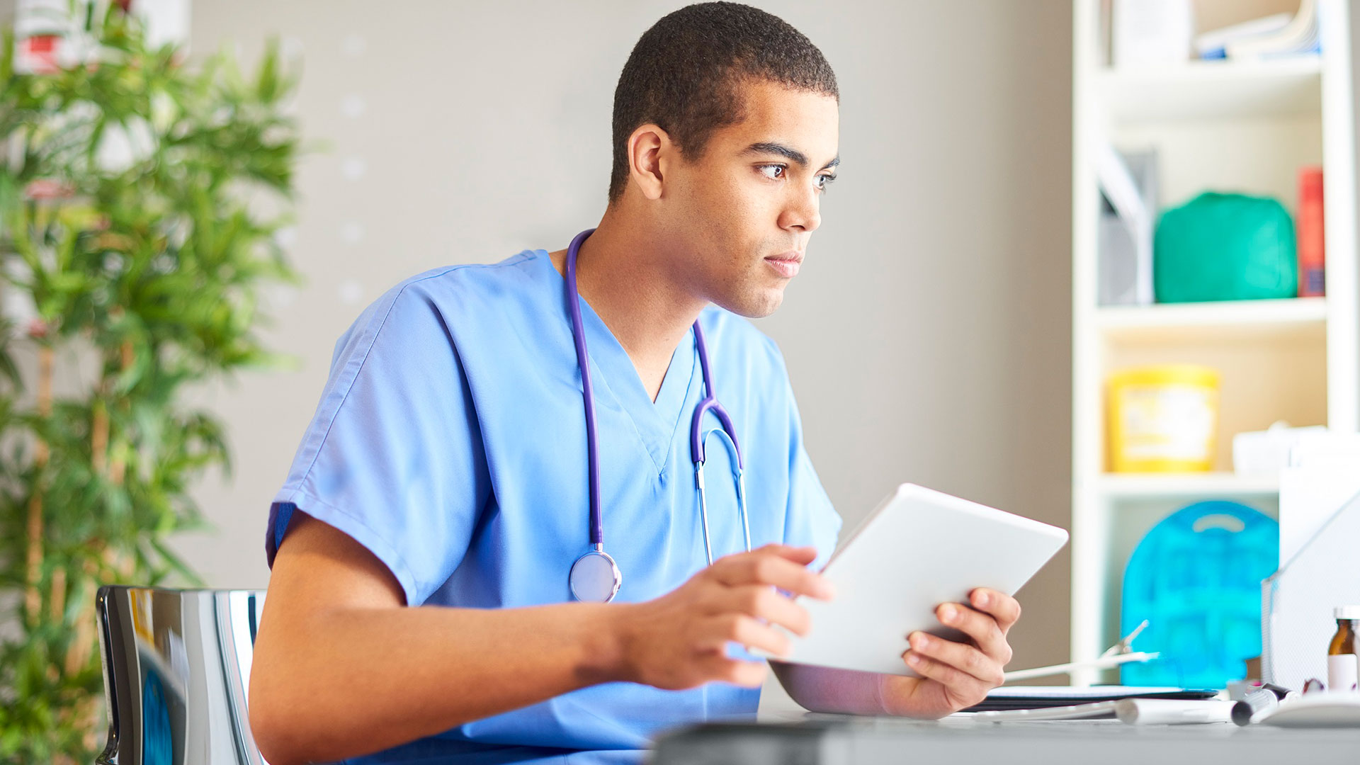 A medical student wearing scrubs. They are sat a desk whilst holding an iPad.
