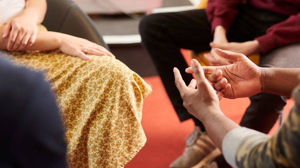 A group counselling and therapy session. There are people sat in a circle talking to each other. Only their hands are in view.