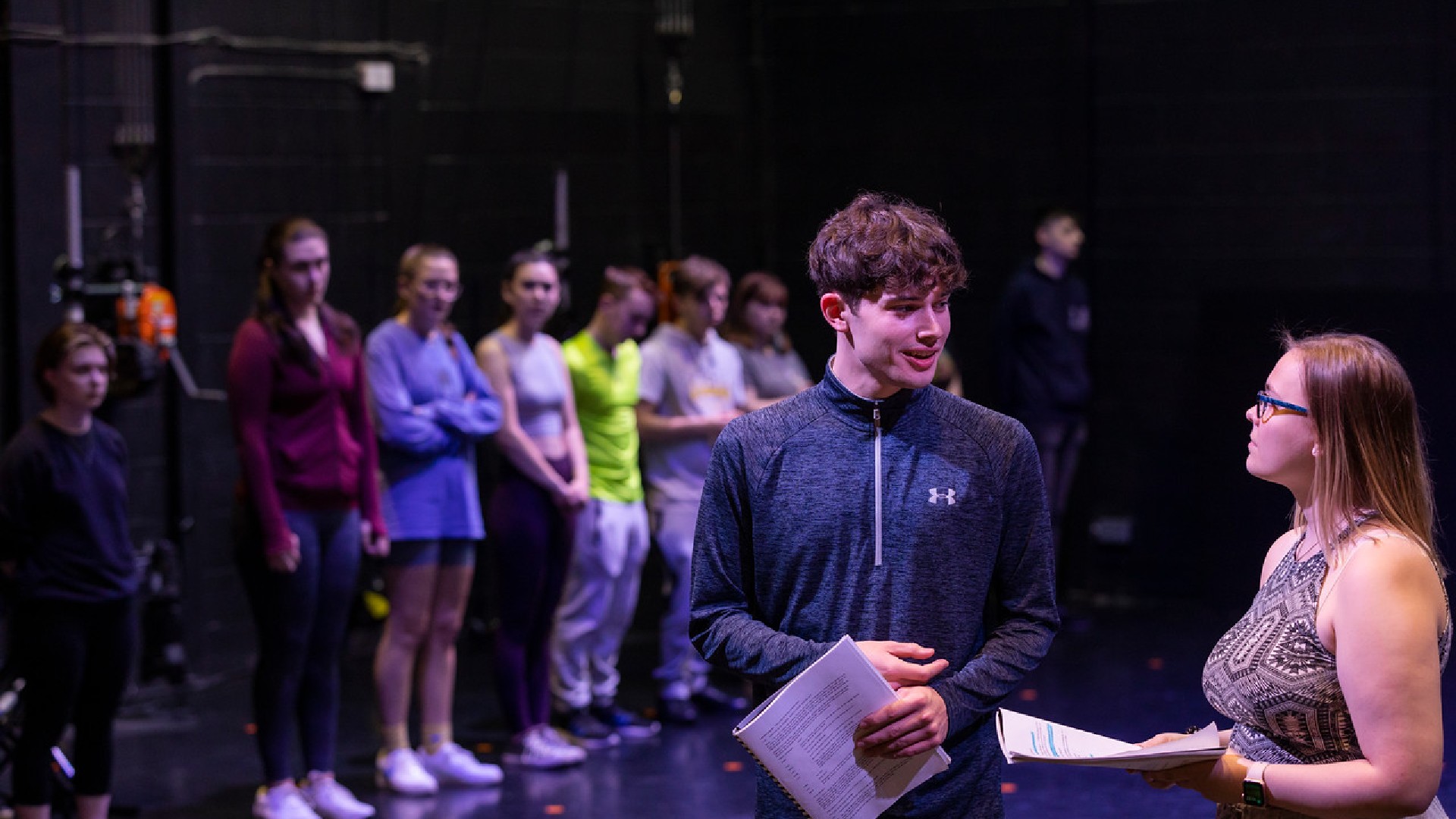 Two people are stood on a stage talking in front of a line of more people.