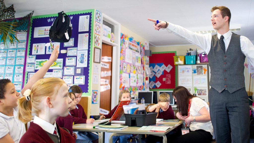A teacher is pointing to a classroom of students. One student has their hand in the air.
