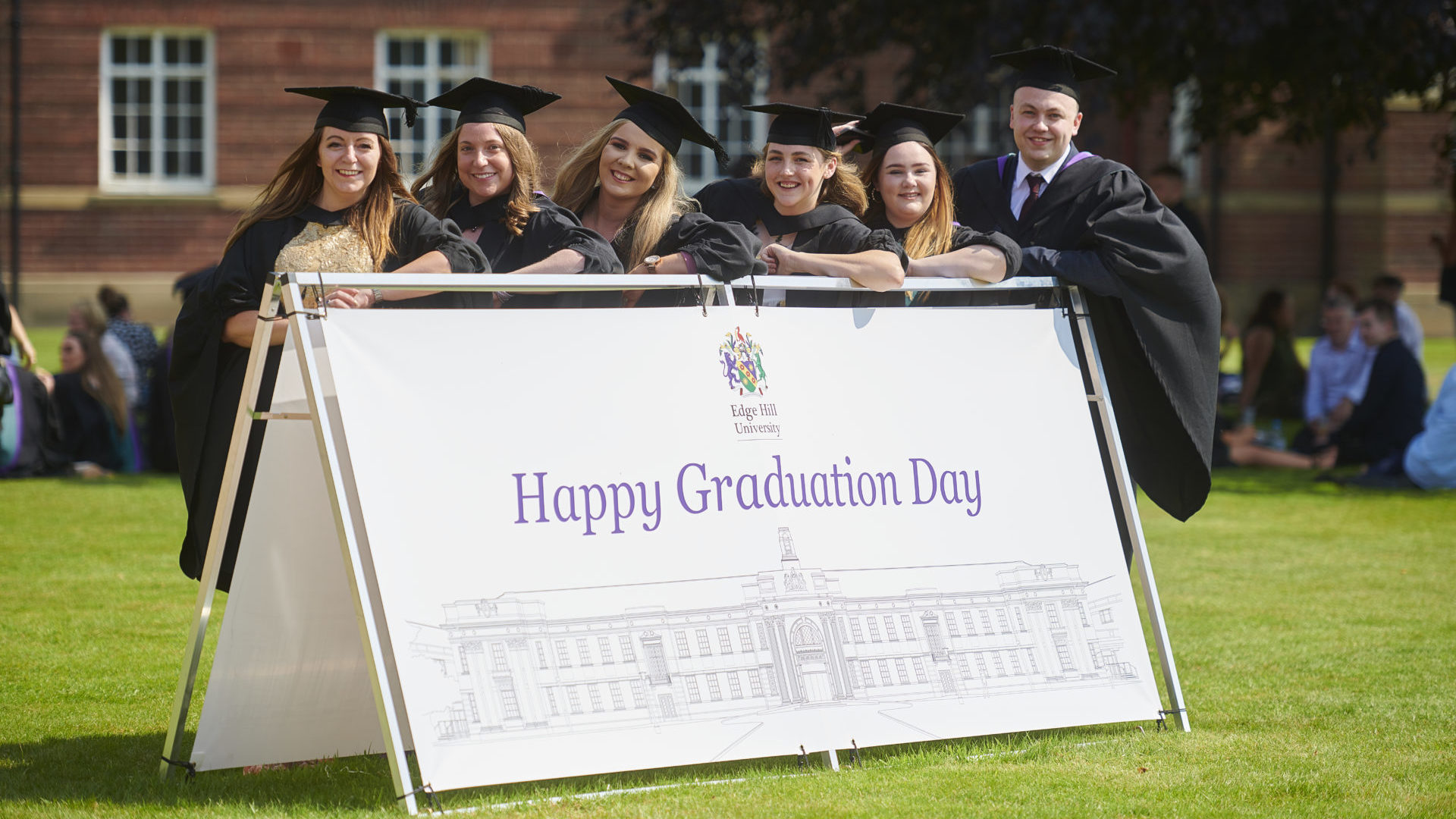 Graduates in front of Happy Graduation Day sign. Clive Myrie James Timpson Sir Terry Leahy Edge Hill graduation.