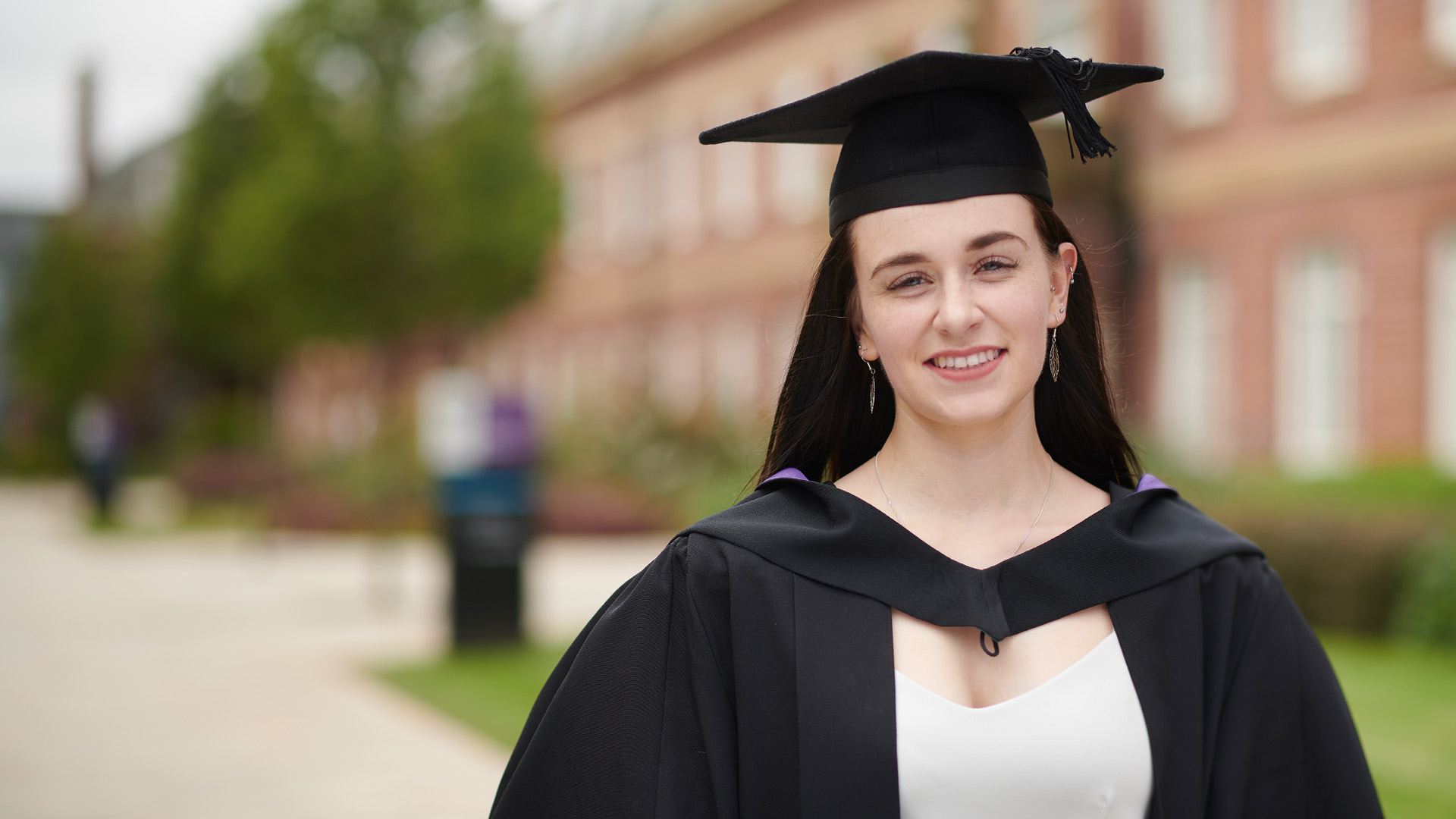 Rachel Redhead wearing a cap and gown and smiling at the camera. Edge Hill University is in the background.