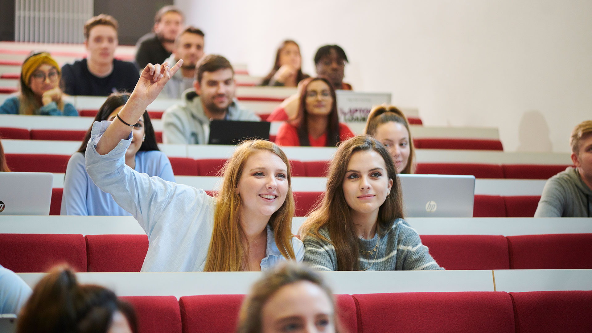 A student is sat in a lecture theatre with their hand up to ask a question during the lecture.