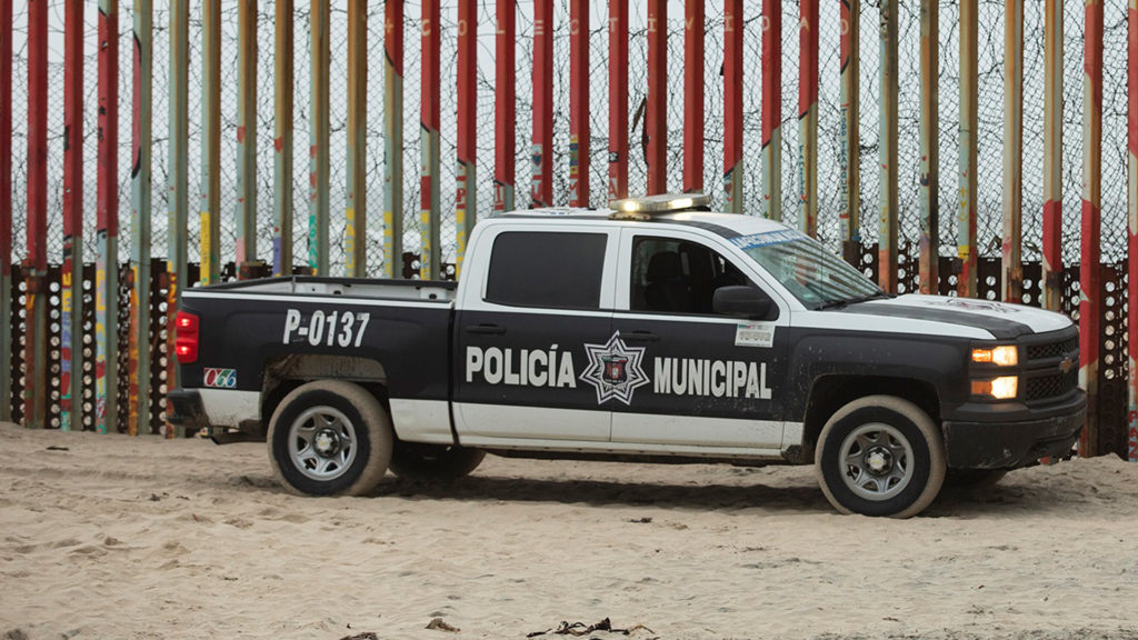 A Mexican police car on sand with a rail and barbed wire fence behind.