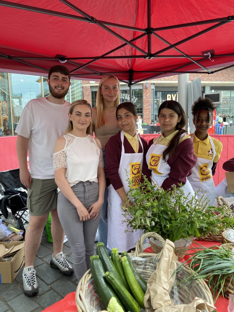 Three students gather with three schoolchildren wearing aprons behind their market stall in Liverpool ONE.