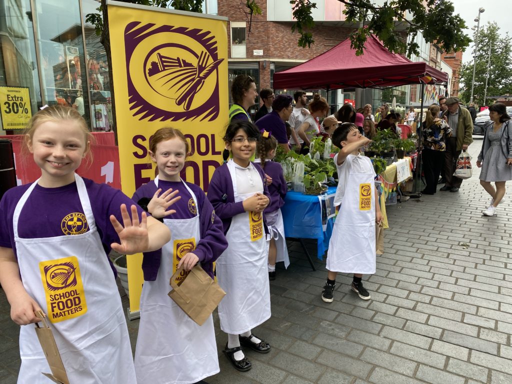 Schoolchildren wearing aprons beckon shoppers over to their market stall.