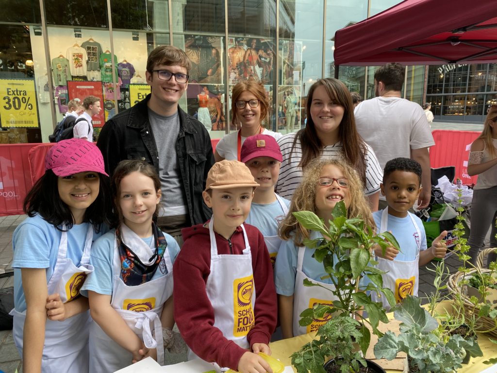 Schoolchildren and teachers stand behind a market stall displaying veg and herbs plants.