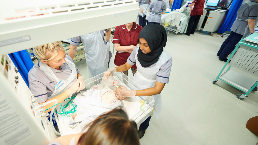 Midwifery students looking into a medical crib wearing gloves and aprons