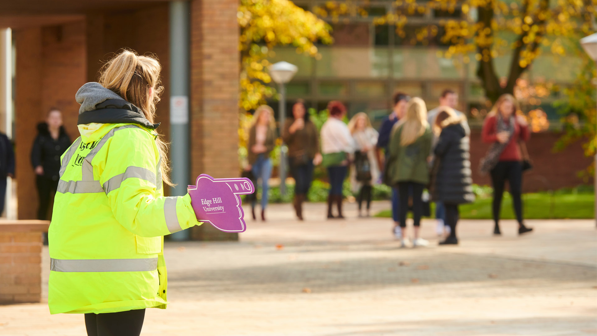 A woman seen from behind in the left of the image, wearing high vis and a purple foam finger that says edge hill university. In the background are a group walking toward her.