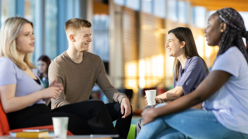 Four students talking on campus holding cups of coffee