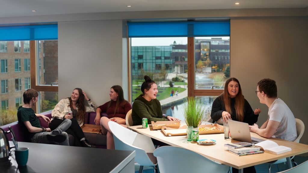 A group of students in the kitchen area of their on-campus accommodation. Three of the students are sitting at the table talking and laughing. Three other students are sitting on the couch in front of the windows.