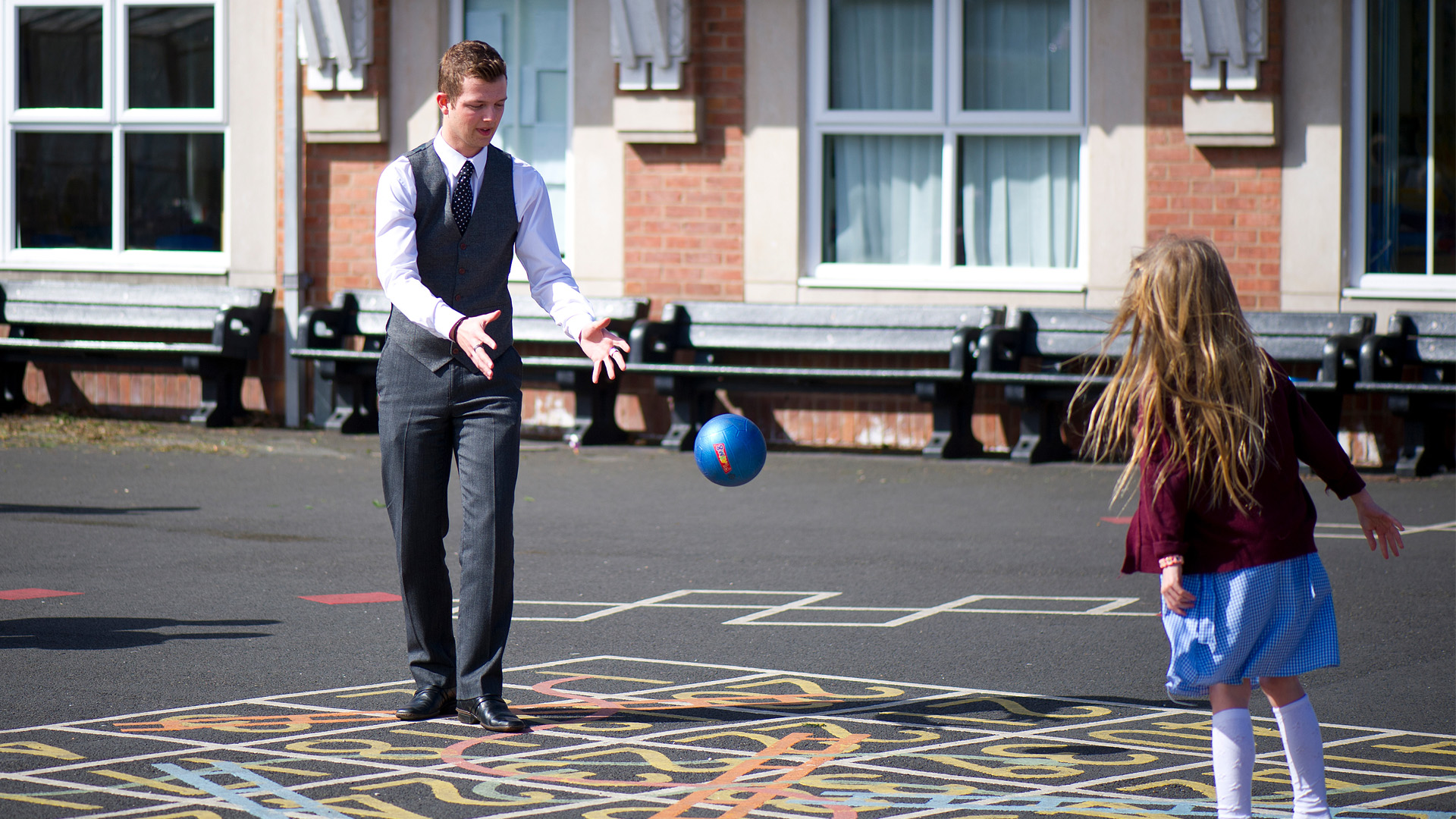 A teacher in a suit and a child with long hair playing with a ball in a playground