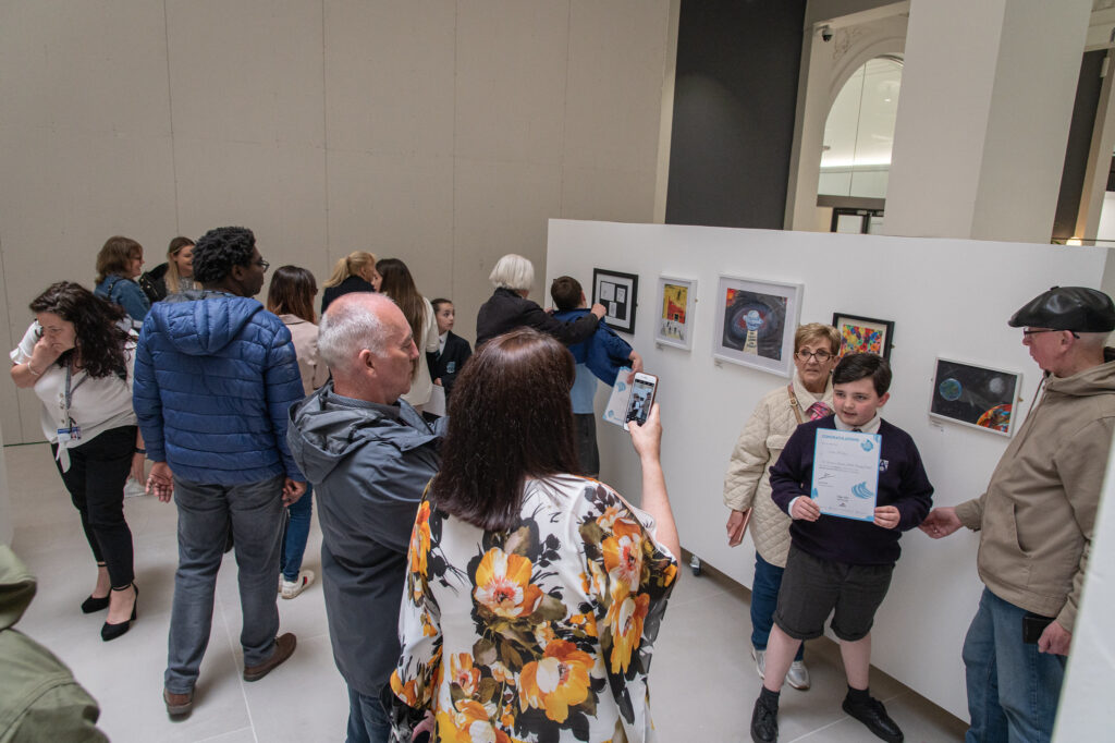 An picture showing the dot-art schools exhibition. In the foreground are parents and guardians taking pictures with children holding certificates. 