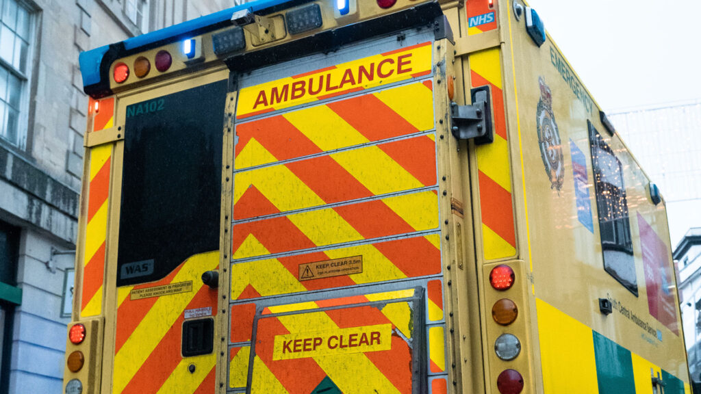 A close up shot of the back of an ambulance. The ambulance is stationary in a city street with its blue lights on.