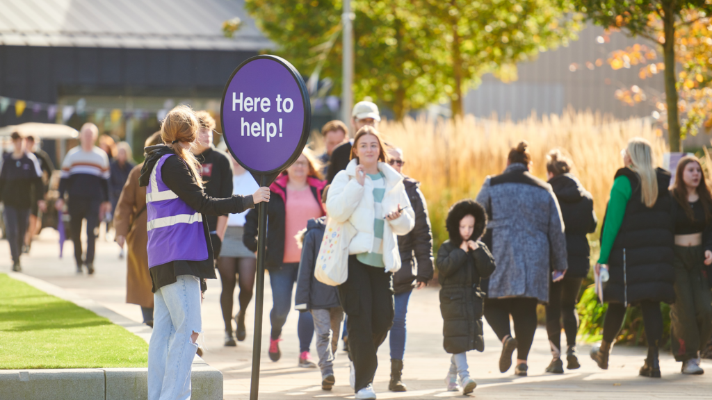 An Edge Hill University student guide holding a sign which reads 'here to help' as a crowd walks past down a busy pathway at a recent open day.