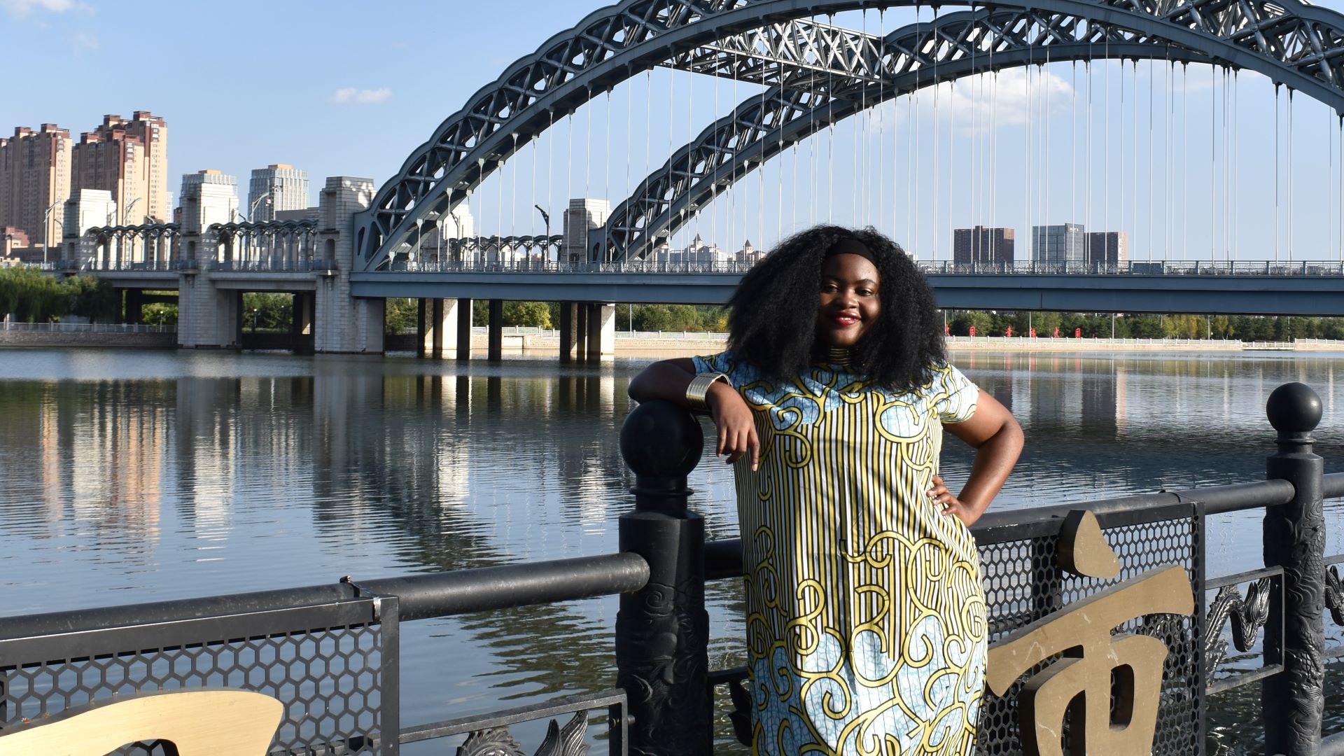 Frances Agba wearing a colourful dress and standing on a bridge.