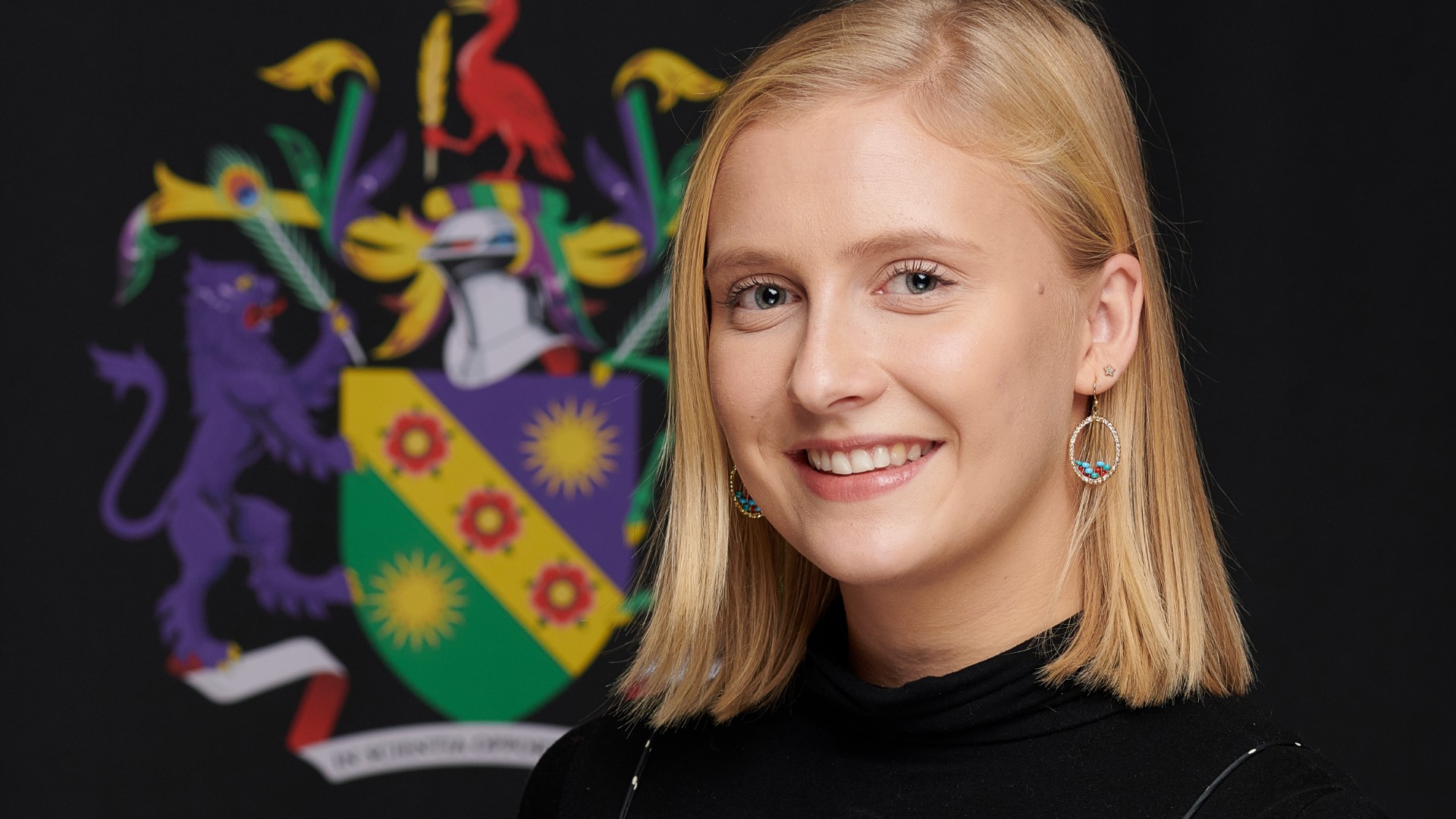 Lydia Horne smiles while standing next to the Edge Hill logo on a black backdrop