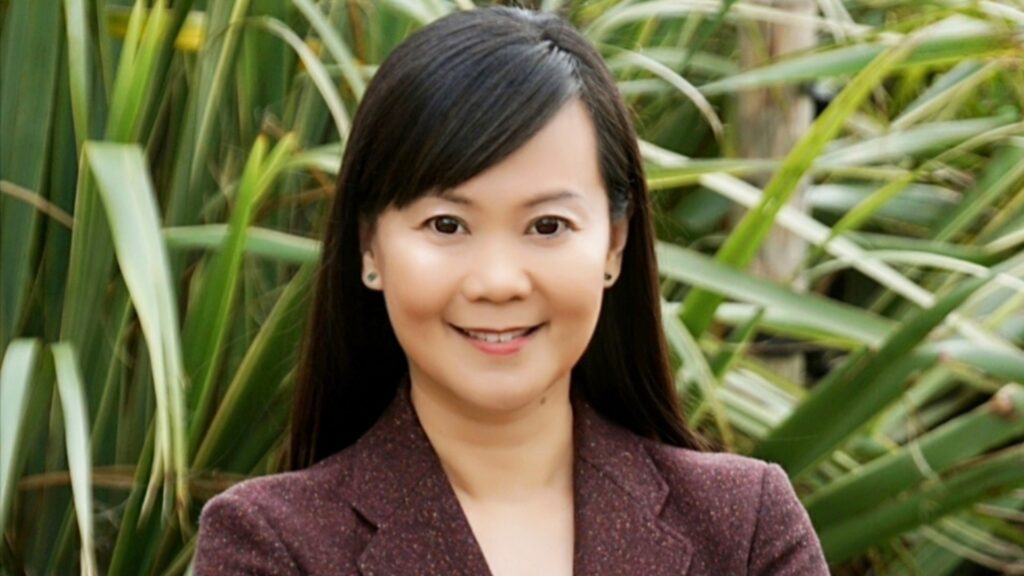 Headshot of Professor May Ng OBE. She is stood in front of palm plants, wearing a maroon blazer and smiling.