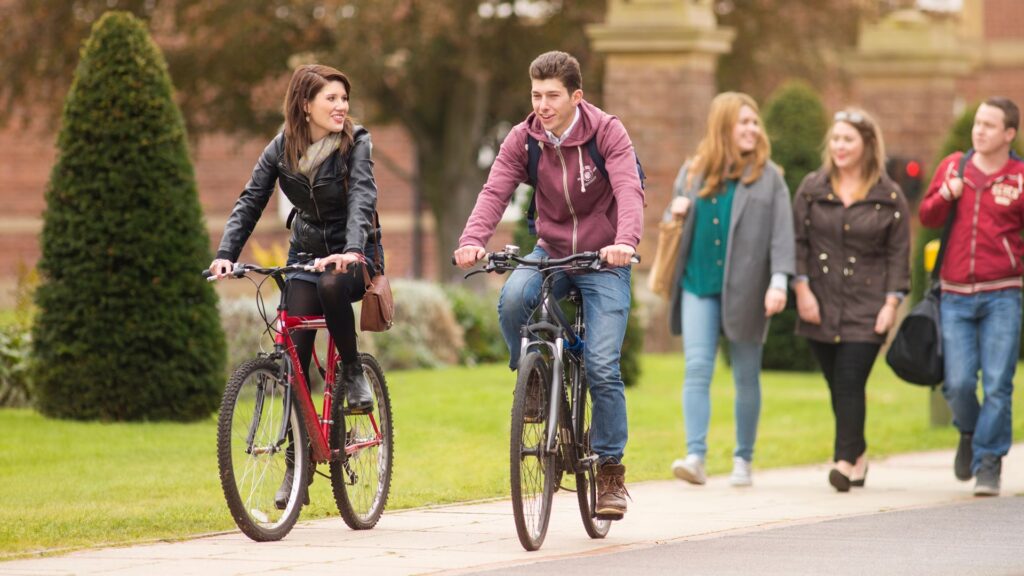 Two students on bikes talking, with some students walking behind near the Main Building