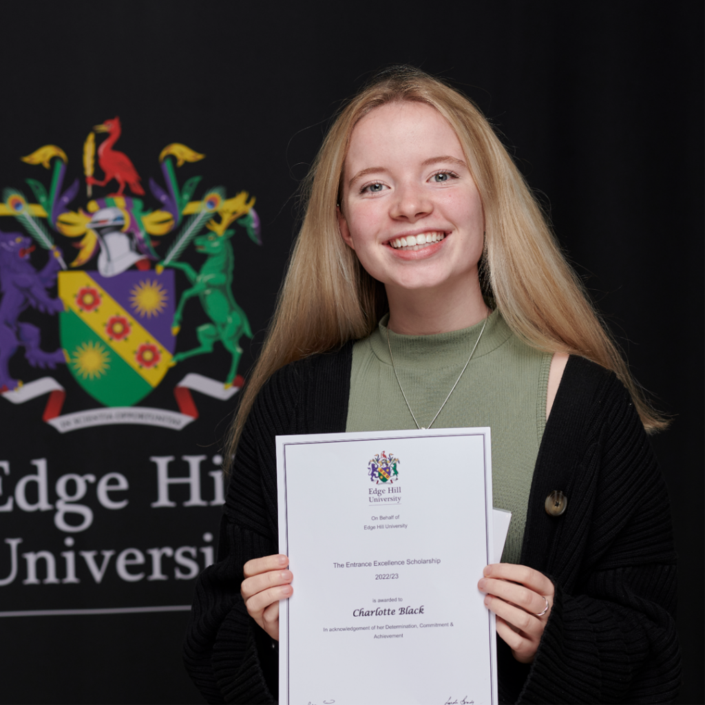 A smiling Charlotte Black holds her Scholarship Award certificate next to the Edge Hill University crest in front of a black background