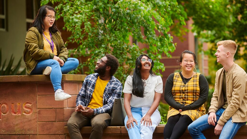 A diverse group of students sit together on a wall on EHU's campus. They are smiling and laughing.