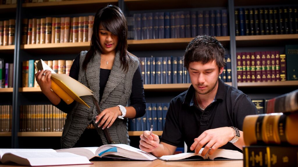 Two student, one stood, one sitting, look through law books whilst stood in front of a large bookshelf
