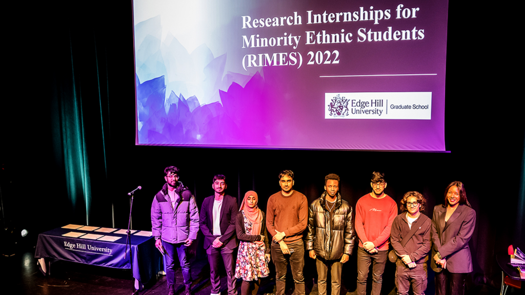 Some of the RIMES interns stand on stage. Above them is a screen displaying the Edge Hill University logo and the text Research Internships for Minority Ethnic Students (RIMES) 2022