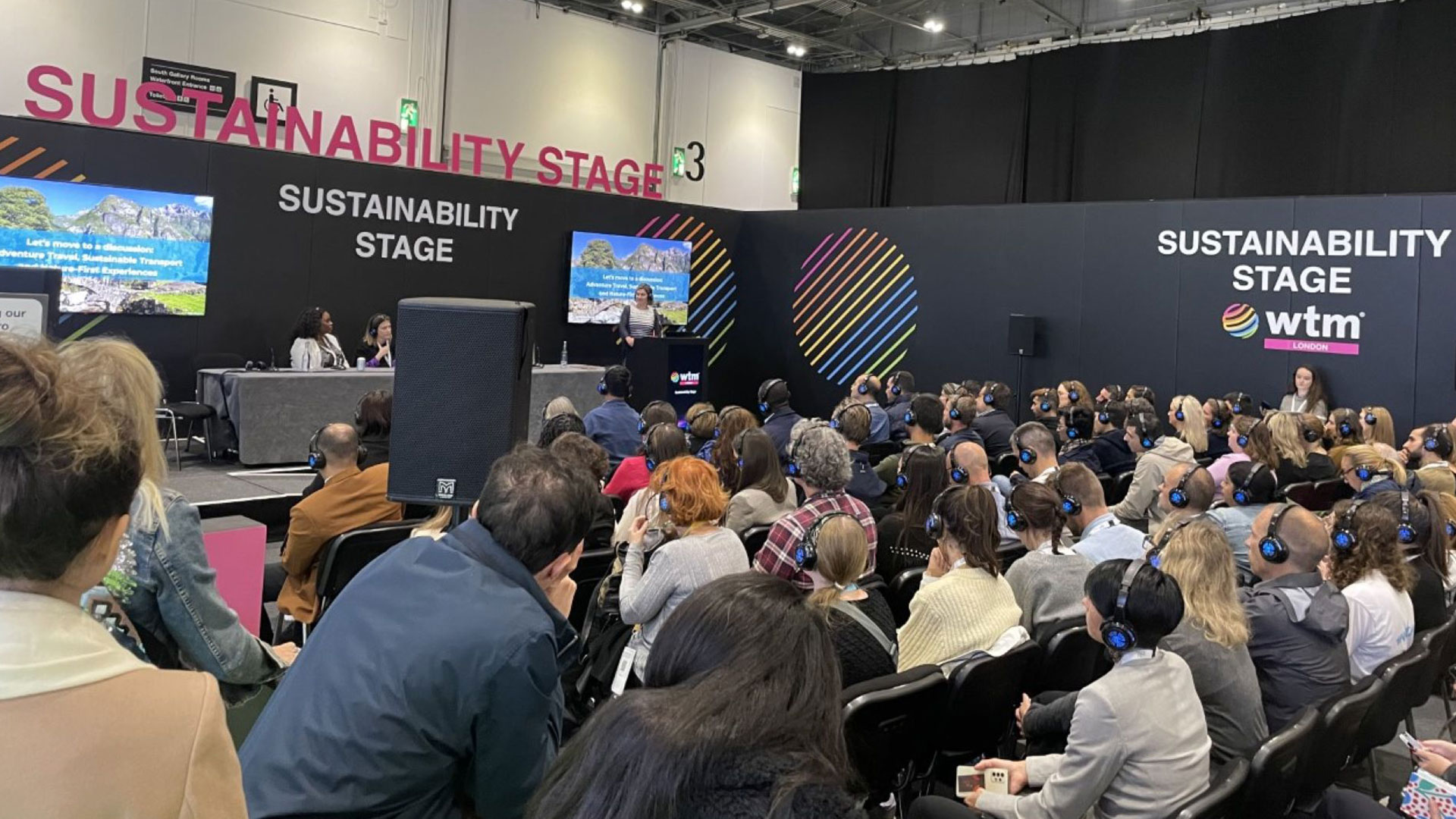 Sustainability stage at the World Travel Market Conference. There is a panel on the stage and the audience are sat in rows, all wearing a headset.