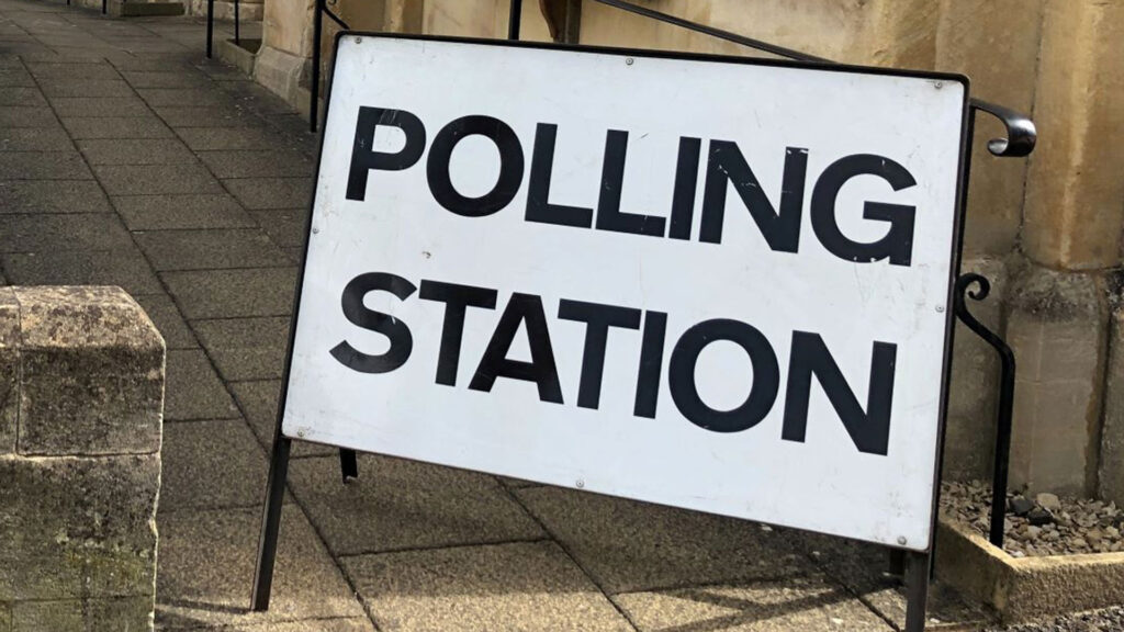 A white sign that reads 'Polling station' in large black font is placed on the pavement in a street to indicate the polling station location