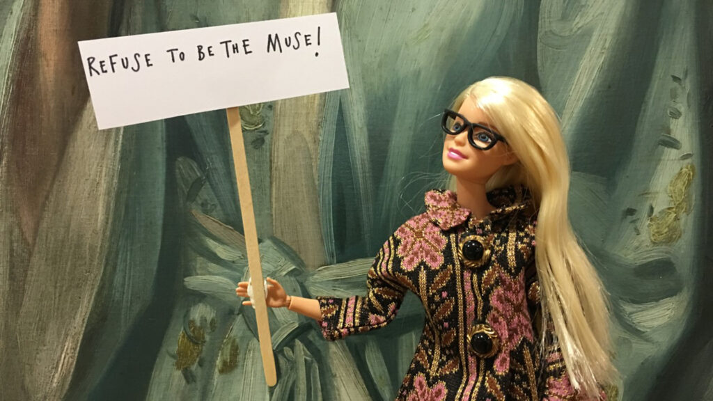 A barbie doll in front of a painting holding a sign saying 'I refuse to be your muse'
