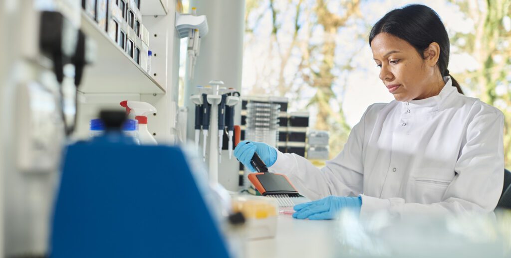 A student using lab equipment in a campus lab. The student is wearing a lab coat and blue gloves.