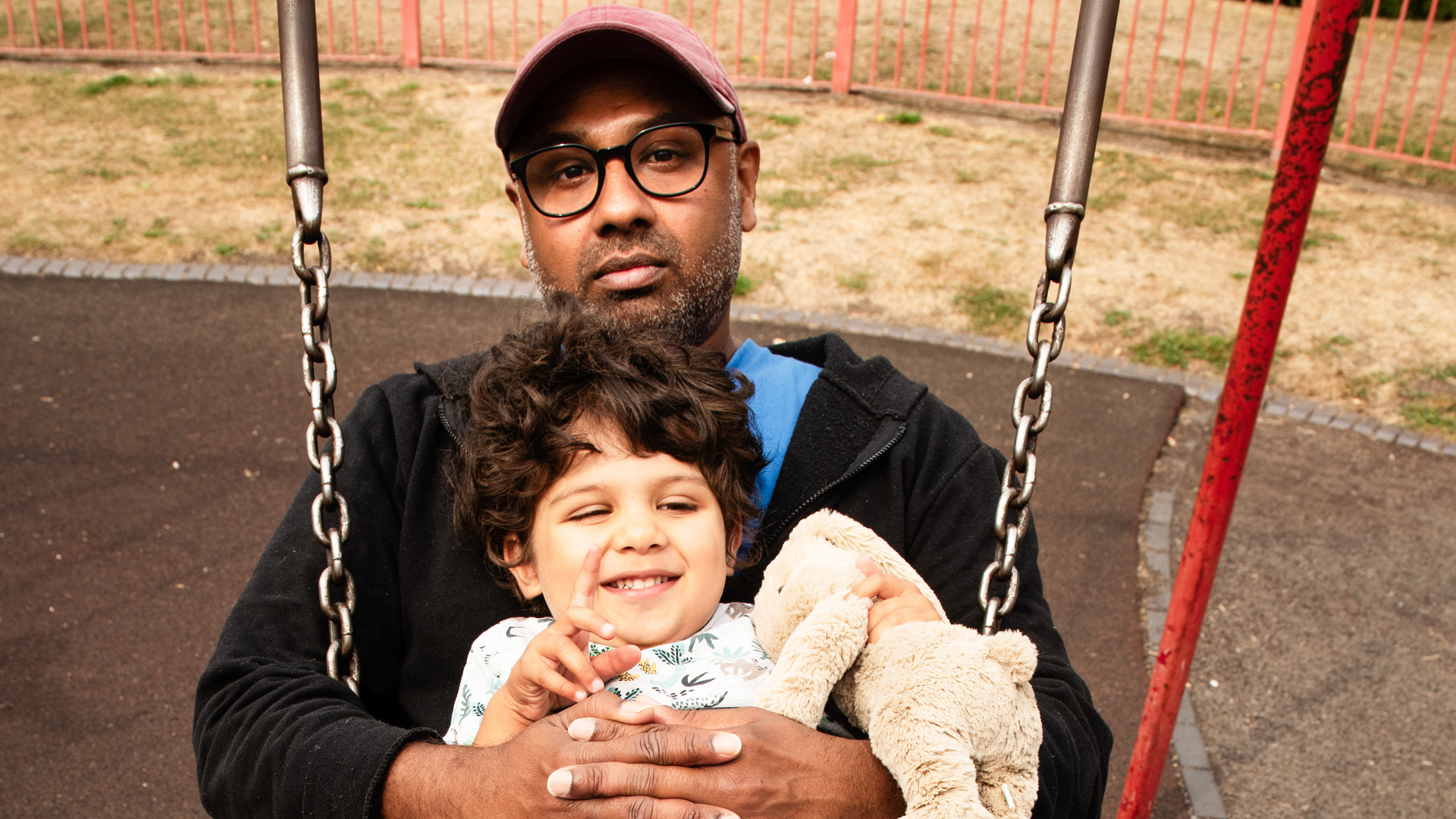 A father sits on a swing, holding their child on their lap.