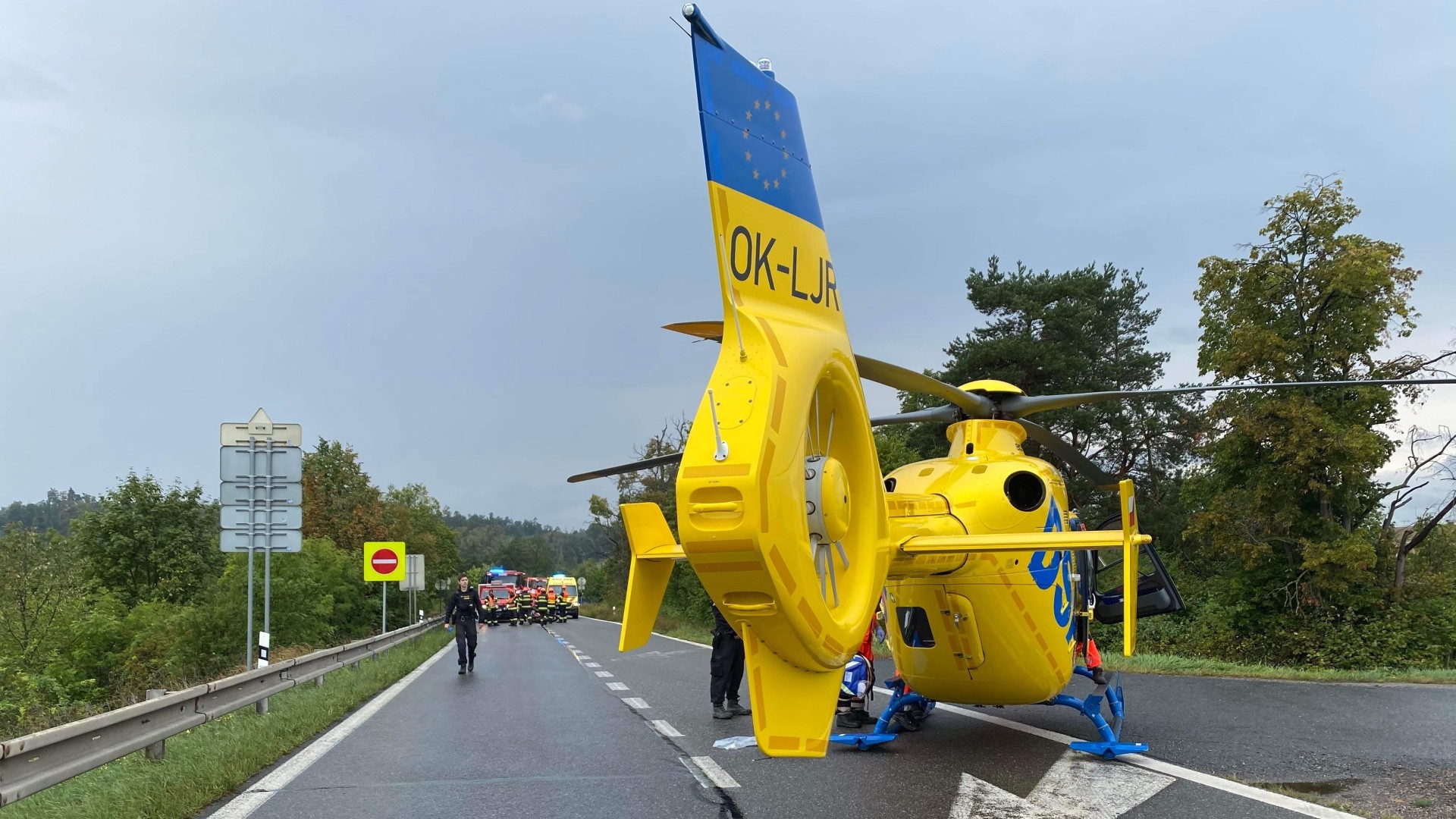 A yellow helicopter sits on a road and a group of emergency services workers stands in the background.