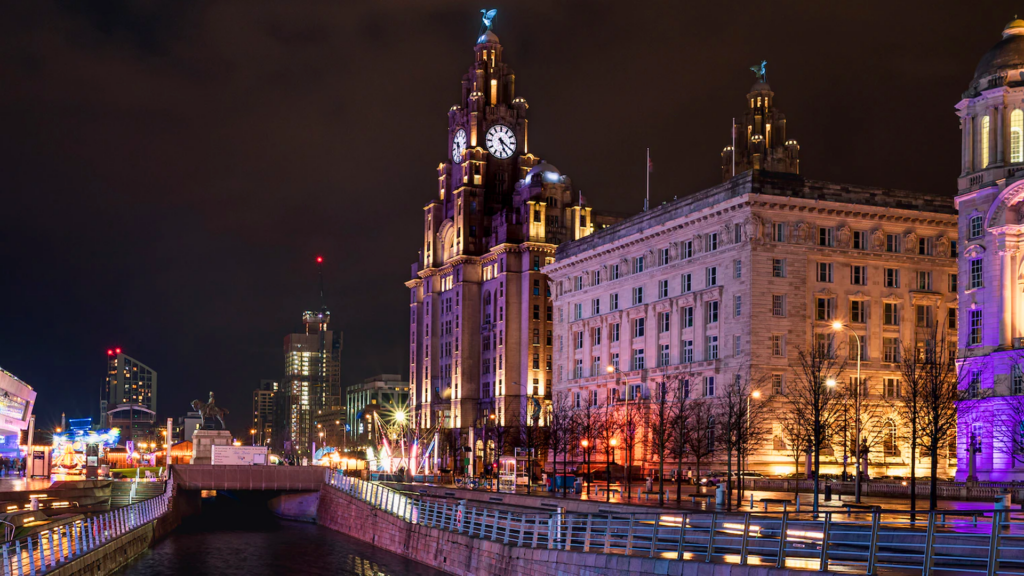 A night-time view of the Royal Liver Buildings in Liverpool City Centre. The buildings are lit up by varicoloured lighting.