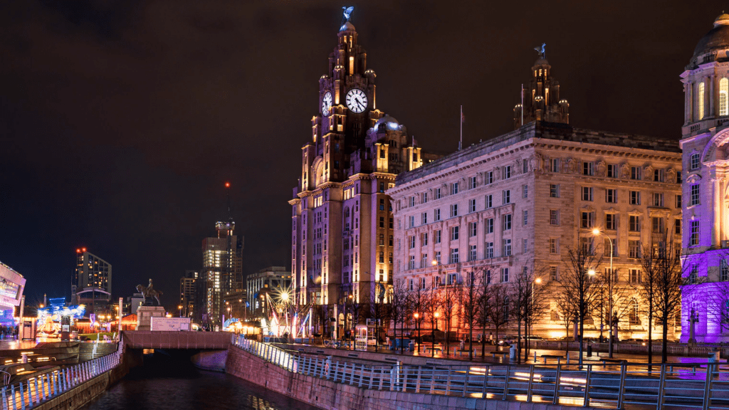 A night-time view of the Royal Liver Buildings in Liverpool City Centre. The buildings are lit up by varicoloured lighting.