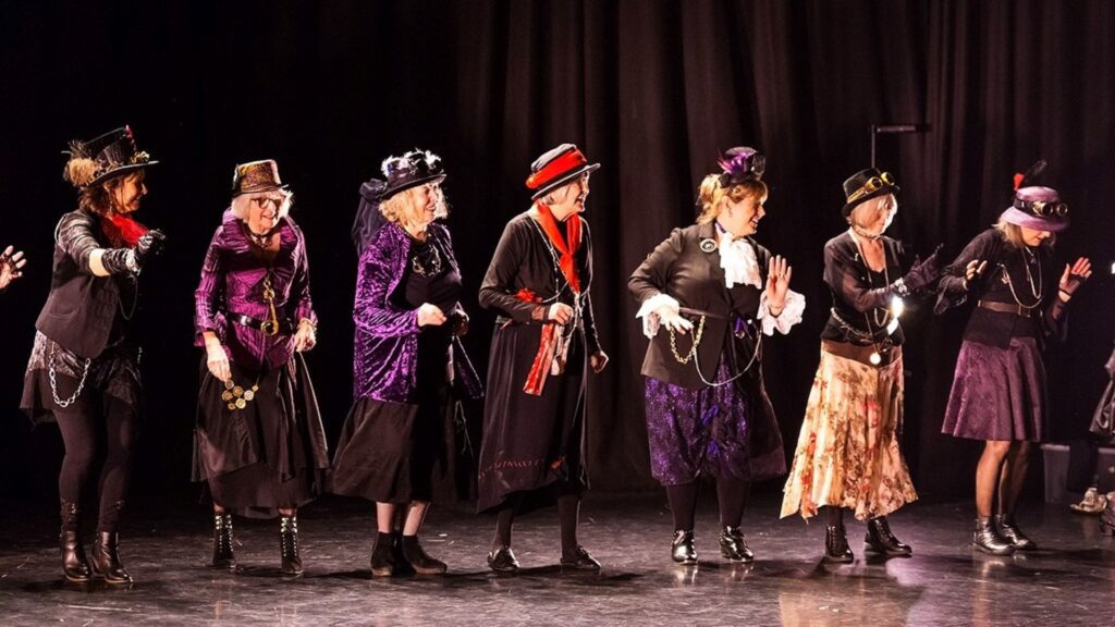 A line of 50 Moves women dancers wearing dresses, jackets and hats dance across a stage as part of the Healthy Brain showcase.