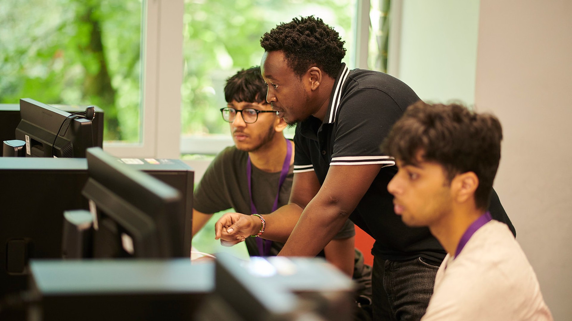 An academic member of staff is stood between two students in an IT suite classroom. The students are using computers whilst the tutor talks to them.