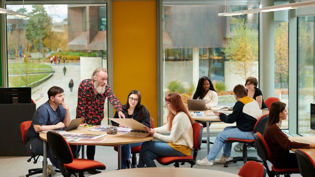 An academic member of staff is teaching students in a classroom. The students are sat around a circle table whilst the academic is talking with them and discussing their work.