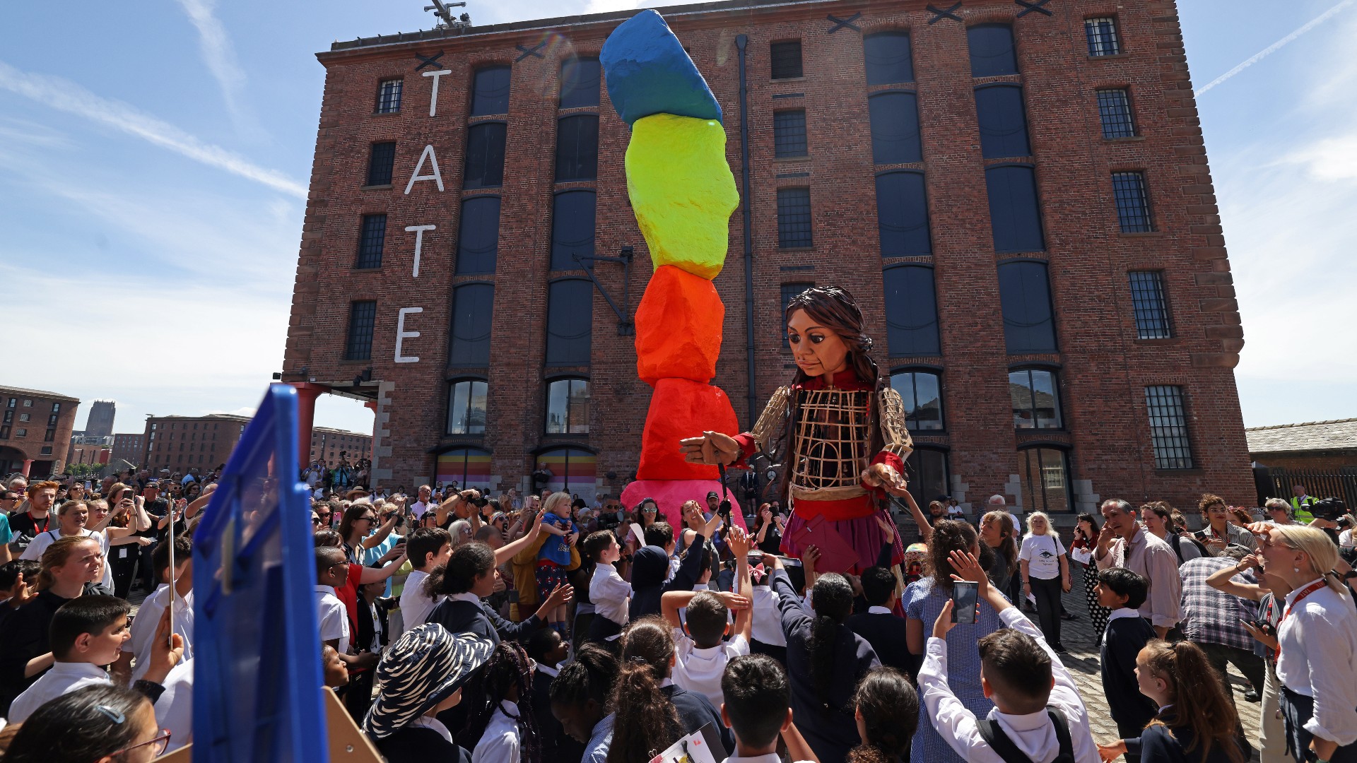 Giant puppet Little Amal stands next to the Liverpool Mountain sculpture surrounded by children from Smithdown Primary School in front of Tate Liverpool.