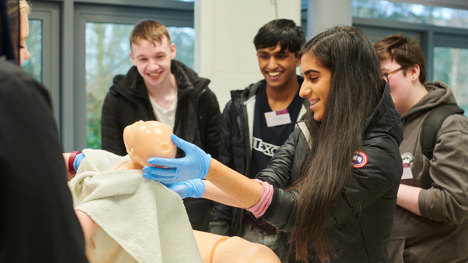 A group of students gather around a simulation patient in bed while one student holds up a and simulation baby.