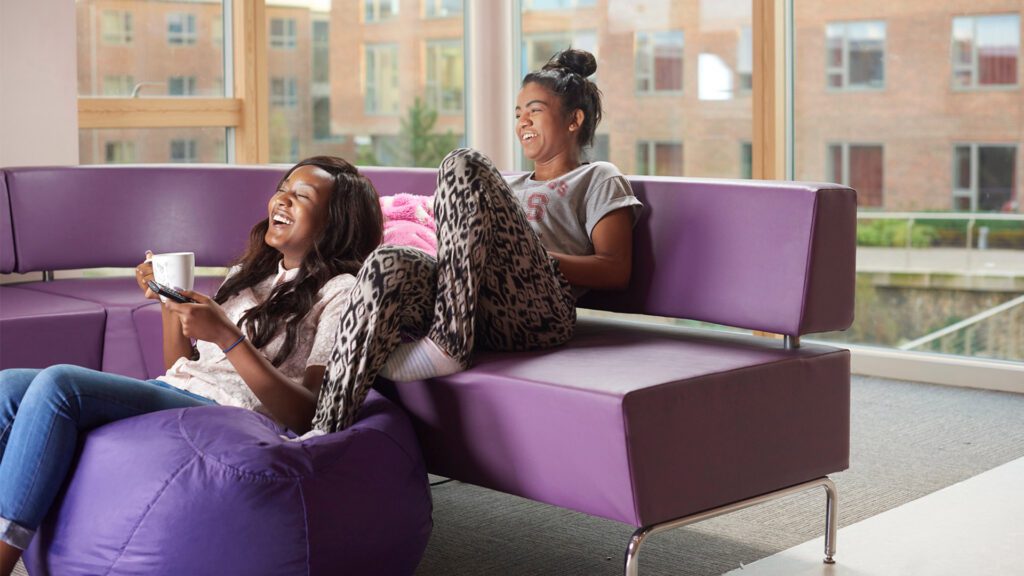 Two students sat on a couch in halls watching tv and laughing