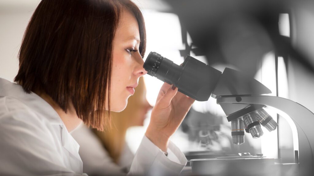 A MEng (Hons) Biomedical Engineering student in white overalls looking down a microscope