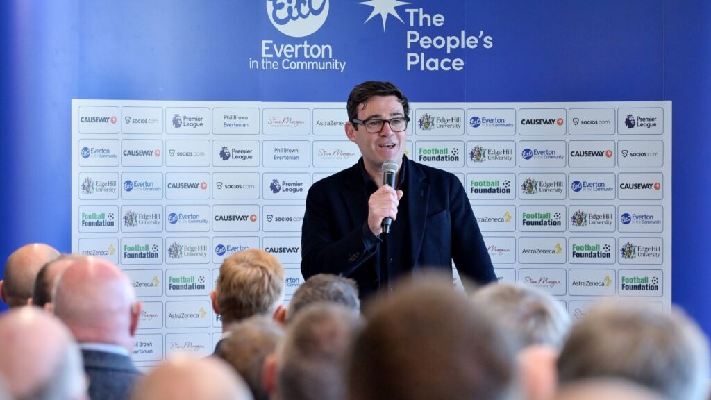 Mayor of Greater Manchester Andy Burnham talks into a microphone with the People's Place sponsorship board behind him and an audience in front.