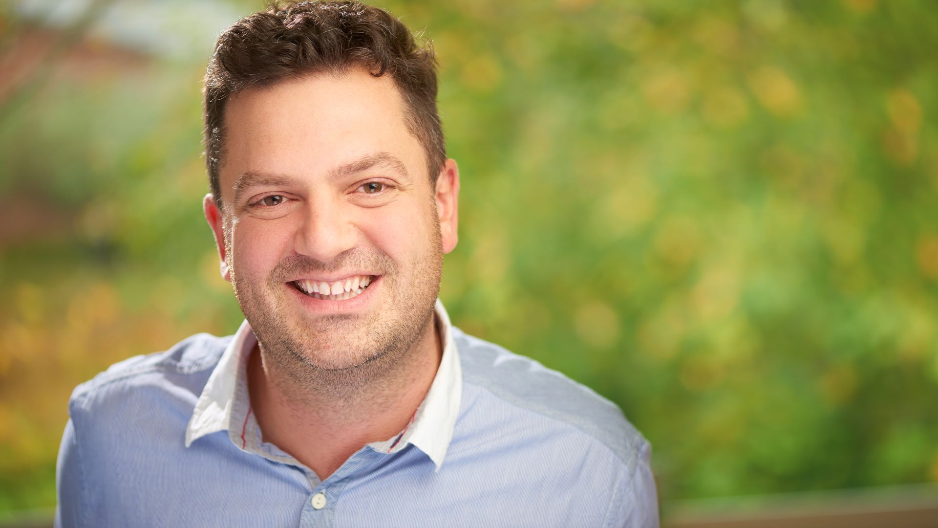 Professor Derek Heim smiles at the camera with a green background behind him.