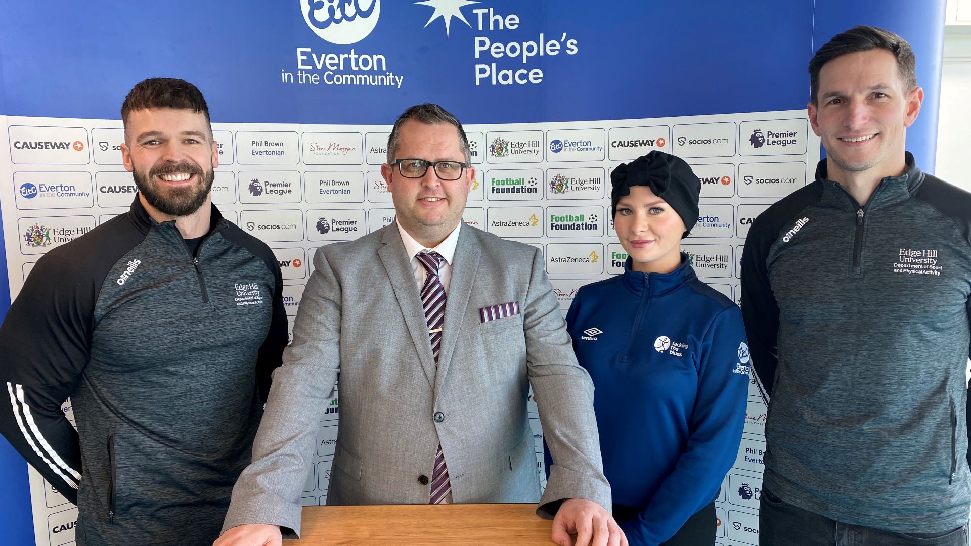 Four people stand in a row with a sponsorship banner behind them at the opening of The People's Place.