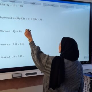 Secondary Mathematics Education with QTS student, Hafsah Nakhuda, completing maths sums on an interactive whiteboard at the front of a classroom.