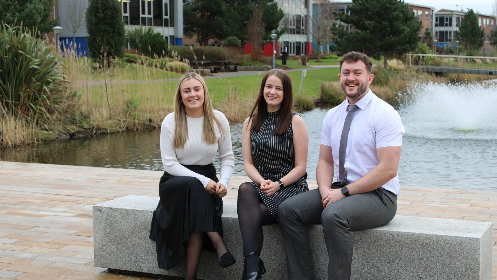 Three smartly dressed students sitting with water behind them.