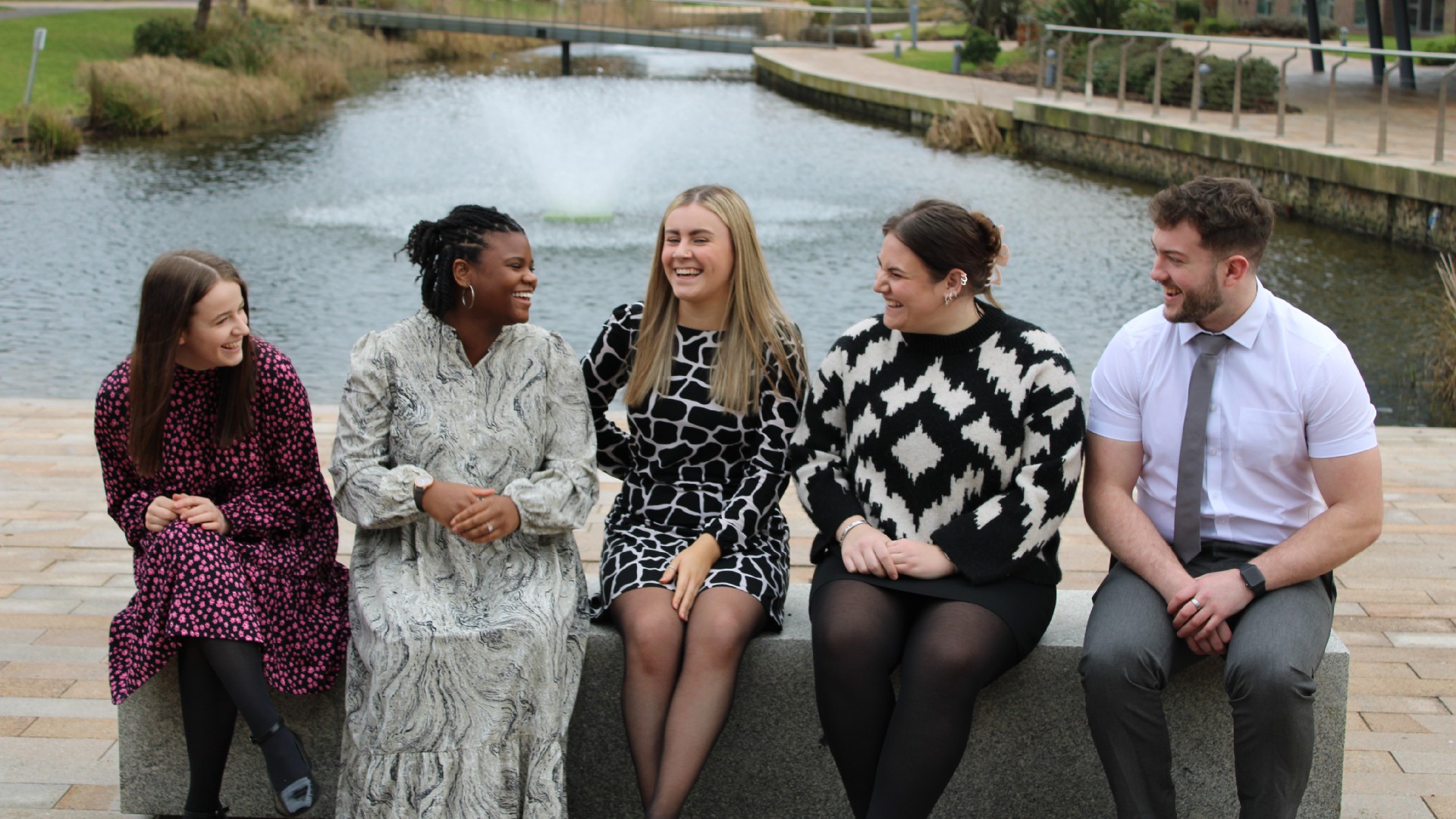 A group of students sitting in front of a lake in campus. The students are all wearing interview clothes.