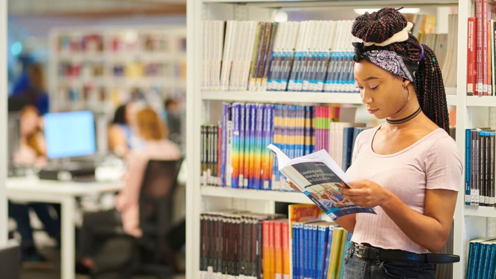 A student stand amongst Catalyst bookshelves, reading a textbook. Their hair is in colourful braids and they are wearing a lilac cropped t-shirt and jeans.