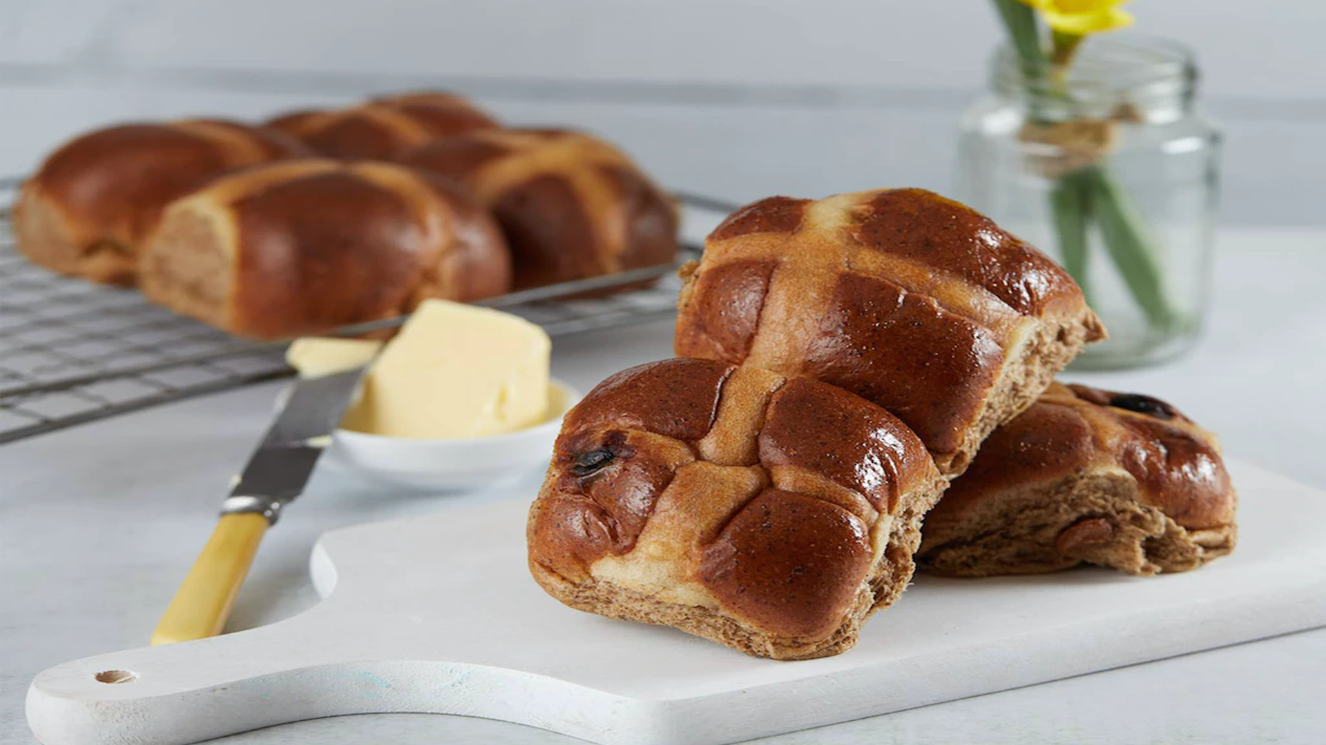 Fresh hot cross buns on a white tray. There is a buttered knife next to them, and a glass vase filled with daffodils.